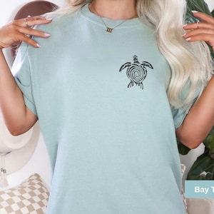 Puede incluir: Camiseta azul claro con un diseño de tortuga negra en la parte delantera. El diseño de la tortuga es un patrón en espiral. La camiseta es de corte holgado. El texto "Bay Tshirt" está en la esquina inferior derecha.