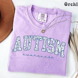 May include: A light purple t-shirt with the word "AUTISM" in large, blue-outlined letters filled with a floral pattern. The word "awareness" is written below in a script font. A silver necklace with a pendant is on the shirt.