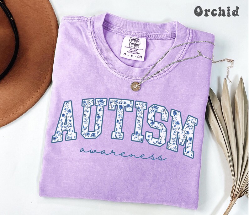 May include: A light purple t-shirt with the word "AUTISM" in large, blue-outlined letters filled with a floral pattern. The word "awareness" is written below in a script font. A silver necklace with a pendant is on the shirt.