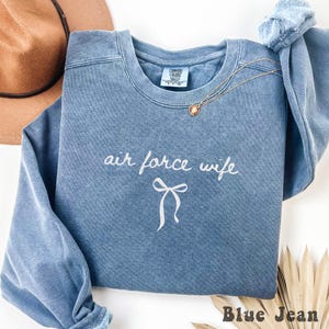 May include: A blue jean-colored sweatshirt with the words "air force wife" in white script and a bow graphic. A gold necklace and a brown hat are visible in the background. The sweatshirt is made of a soft material.