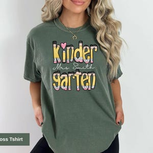 May include: Moss green t-shirt with the words "Kindergarten Mrs. Smith" in a colorful, pencil-themed design. The shirt features a heart above the word "Kinder". The shirt is a comfortable, casual style.