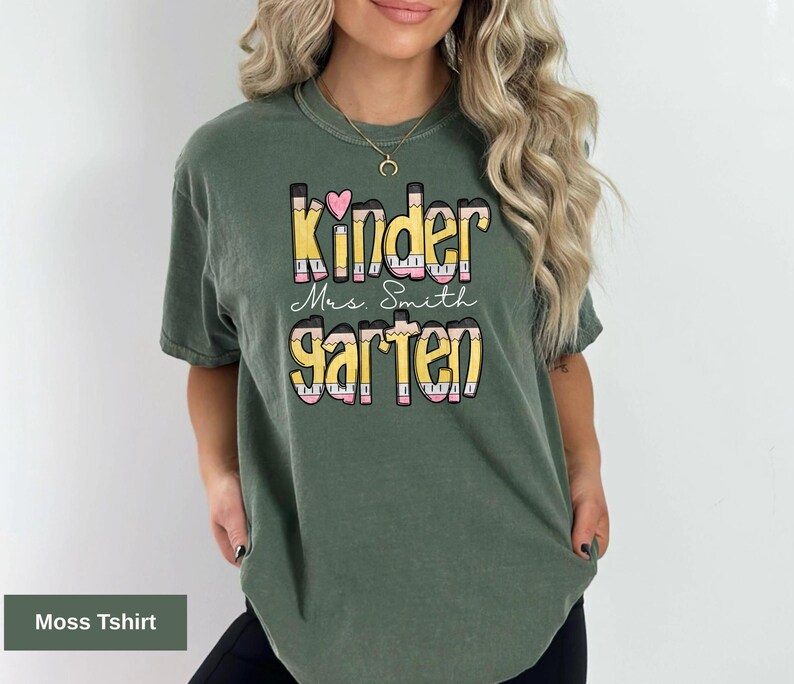 May include: Moss green t-shirt with the words "Kindergarten Mrs. Smith" in a colorful, pencil-themed design. The shirt features a heart above the word "Kinder". The shirt is a comfortable, casual style.
