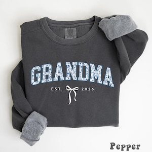 May include: A dark gray sweatshirt with the word "GRANDMA" in blue floral print. Below the word "GRANDMA" is the text "EST. 2026" with a white bow graphic. The sweatshirt has gray fleece lining the cuffs.