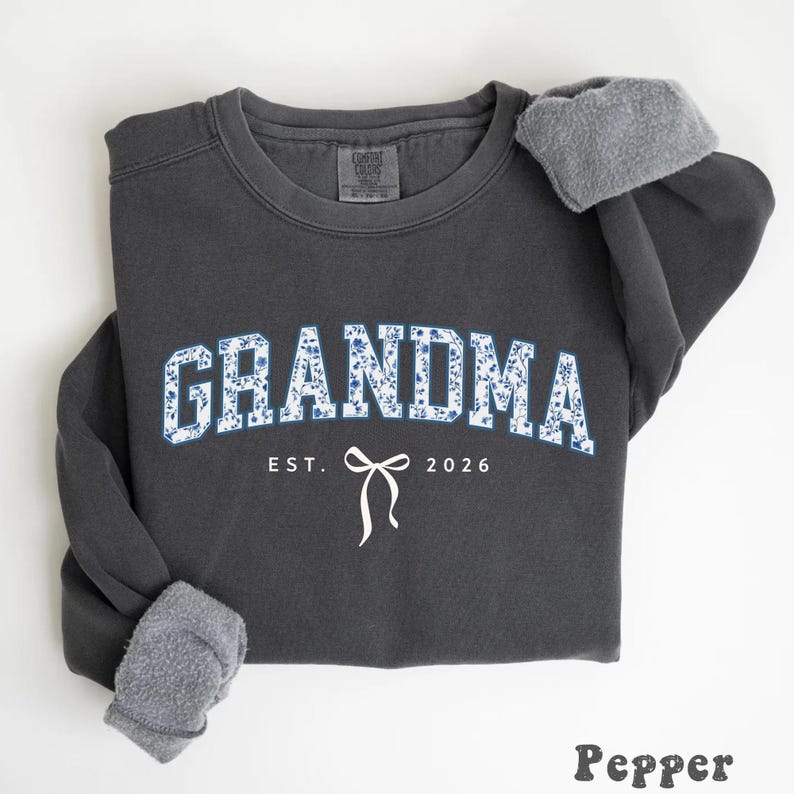 May include: A dark gray sweatshirt with the word "GRANDMA" in blue floral print. Below the word "GRANDMA" is the text "EST. 2026" with a white bow graphic. The sweatshirt has gray fleece lining the cuffs.