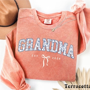 May include: Terracotta-colored sweatshirt with the word "GRANDMA" in blue floral print lettering. Below the word "GRANDMA" is the text "EST. 2026" and a white bow graphic. A brown hat and gold necklace are visible.