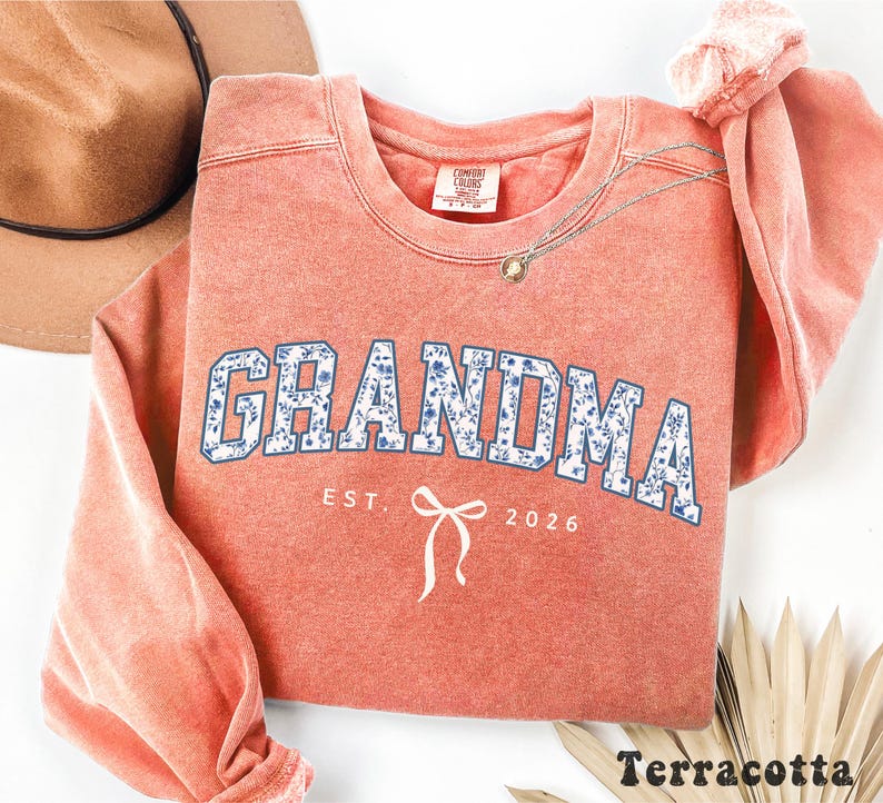May include: Terracotta-colored sweatshirt with the word "GRANDMA" in blue floral print lettering. Below the word "GRANDMA" is the text "EST. 2026" and a white bow graphic. A brown hat and gold necklace are visible.