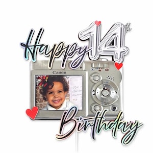 May include: A silver camera-shaped cake topper with the words "Happy Birthday" and "14th" in a holographic font. The camera displays a photo of a smiling child and is decorated with red heart accents.