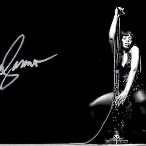 Puede incluir: Fotografía en blanco y negro de una cantante en el escenario, sosteniendo un pie de micrófono. La cantante lleva un vestido con lentejuelas. La imagen incluye una firma en la esquina superior izquierda.