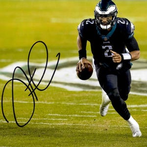 May include: A signed photograph of a football player in a black and teal uniform, running on a green field. The player is holding a brown football and wearing a helmet with a white and teal design. The signature is in black ink.