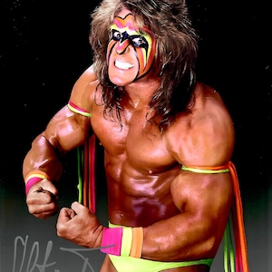 Może przedstawiać: Umięśniony zapaśnik z malowaniem twarzy w żywych kolorach, w neonowych zielonych spodenkach i kolorowych opaskach na nadgarstki i ramiona. Widoczne są również długie, falowane włosy i podpis. Zapaśnik napina mięśnie.