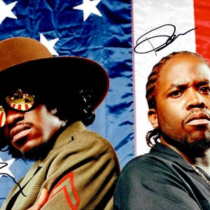 Op de afbeelding: Twee mannen poseren voor een achtergrond van de Amerikaanse vlag. De ene man draagt een bruine hoed, een zonnebril met een rode, witte en blauwe reflectie en een jas in militaire stijl. De andere man draagt een zwart overhemd en een zilveren ketting.