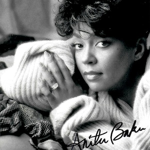 Pode incluir: Retrato a preto e branco de Anita Baker, com a sua assinatura. Ela usa um suéter texturizado e um anel. O seu cabelo escuro está penteado num corte curto e encaracolado. A imagem é um close-up.