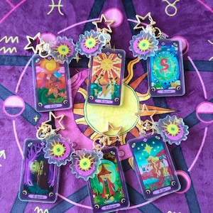 May include: A collection of tarot card keychains featuring colorful illustrations. Each keychain has a gold-colored star and sun charm. The cards display images of The Sun, The Star, The Magician, and other tarot cards. The background is a purple cloth with astrological symbols.