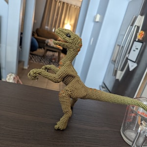 May include: A green crocheted dinosaur toy with orange accents. The dinosaur is standing on a dark brown wooden surface.