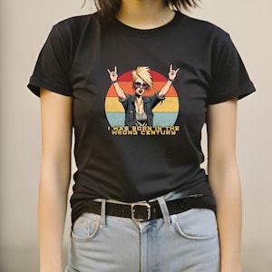 May include: A black t-shirt with a retro-style graphic of a woman with sunglasses and a leather jacket. The graphic includes the text "I was born in the wrong century".