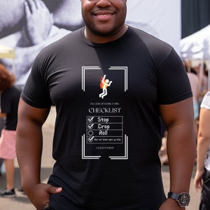 Peut inclure: T-shirt noir avec un graphique blanc d'une personne tombant d'un immeuble en feu avec le texte "IN CASE OF BODILY FIRE CHECKLIST" et une liste de contrôle avec les options "Stop", "Drop", "Roll", et "Shut em' down open up shop". Le texte "#SAFETYFIRST" est en bas du graphique.