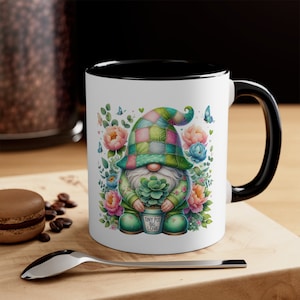 May include: A white ceramic mug with a black rim and handle features a colorful illustration of a gnome holding a potted succulent. The gnome is wearing a green and pink patchwork hat and a green sweater. The text on the pot reads "Tiny Pot Big Love".