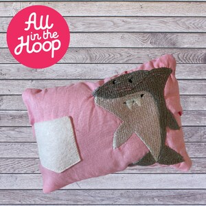 May include: A pink pillow with a white pocket and an embroidered gray shark. The pillow has the text "All in the Hoop" on a red circle.