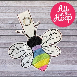 May include: A white leather keychain with a rainbow bee design. The bee has black eyes, a purple and blue stripe, and a rainbow stripe on its abdomen. The keychain has a silver metal ring.