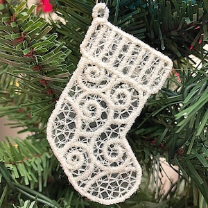 May include: White lace Christmas stocking ornament with a swirl pattern. The stocking is hanging on a green artificial Christmas tree.