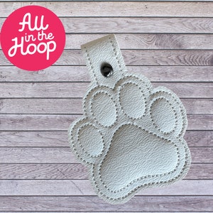 May include: A white paw print keyring with a stitched outline and a press stud closure. The keyring has the text "All in the Hoop" in a pink circle. The background is a light wood grain.