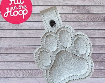 May include: A white paw print keychain with a stitched outline and a snap closure. The keychain has the text "All in the Hoop" in a pink circle. The background is a light wood grain.
