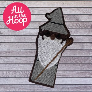 May include: Embroidered patch featuring a wizard with a gray hat, white beard, and a brown staff. The patch has a brown border and is set against a wooden background. The text "All in the Hoop" is in a pink circle.