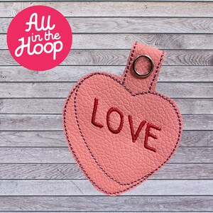 May include: A pink heart-shaped keychain with the word "LOVE" embroidered in red. The keychain has a textured surface and a metal ring for attachment. The text "All in the Hoop" is in a pink circle in the upper left corner.