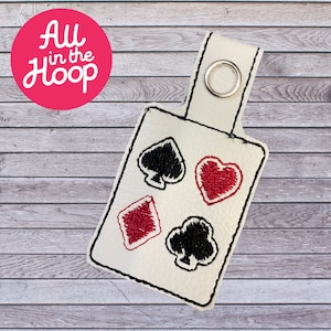 May include: White leather keychain with a black and red embroidered design of playing card suits. The suits are a heart, diamond, and two spades.