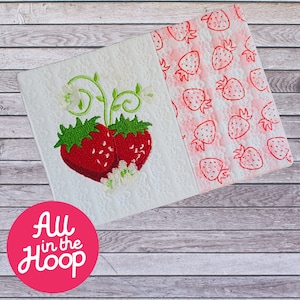 Pode incluir: Uma toalha de cozinha branca com um desenho de morango bordado vermelho de um lado e uma estampa de morango vermelho do outro lado. A toalha está dobrada ao meio e tem o texto "All in the Hoop" em um círculo vermelho.