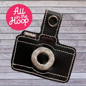 May include: Black leather camera keychain with white stitching and a silver ring. The camera has a white lens and a red detail on the top.