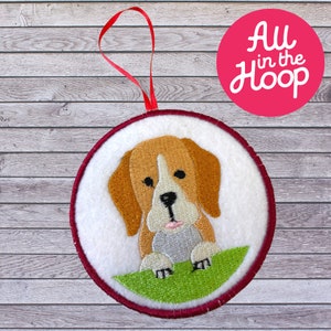 Puede incluir: Un adorno redondo de fieltro blanco con un borde rojo, que presenta un perro beagle bordado con pelaje marrón y blanco, mirando hacia arriba con la lengua rosa sobresaliendo. El adorno tiene un lazo de cinta rojo para colgar.