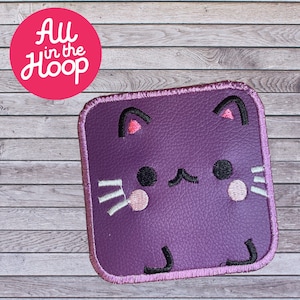 Pode incluir: Patch quadrado de feltro roxo com um rosto de gato de desenho animado bordado. O gato tem orelhas cor-de-rosa, olhos pretos, bigode branco e nariz rosa.