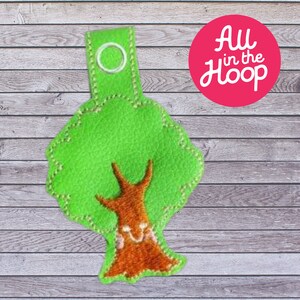 May include: A green felt tree with a brown felt trunk and a smiling face. The tree has a loop at the top for hanging. The text "All in the Hoop" is visible in the background.