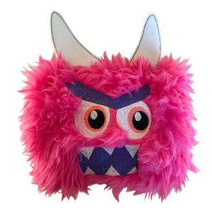 May include: A pink plush monster with white horns, purple eyebrows, and a purple and white mouth. The monster has orange eyes with black pupils.