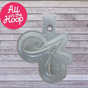 Op de afbeelding: Een zilveren leren sleutelhanger met een decoratief letter "G" ontwerp. De sleutelhanger heeft een lus om aan een tas of sleutels te bevestigen.