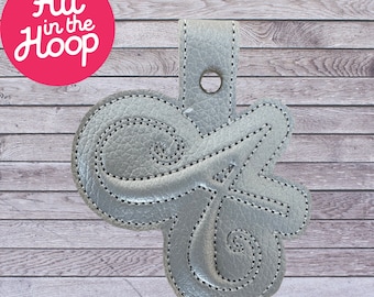 May include: A silver leather keychain with a decorative letter "G" design. The keychain has a loop for attaching to a bag or keys.