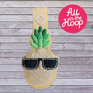 May include: A beige leather keychain with an embroidered pineapple wearing sunglasses. The pineapple has green leaves and a yellow body. The keychain has a metal ring at the top.