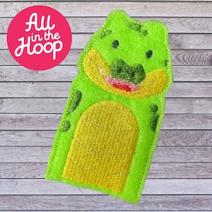 May include: Green felt finger puppet shaped like an alligator with a yellow belly and a red mouth. The puppet is on a wooden background.