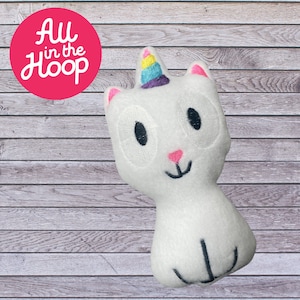 Può includere: Giocattolo di peluche bianco a forma di gatto con un corno di unicorno arcobaleno, occhi ricamati e un naso rosa. Il giocattolo è seduto su una superficie di legno.