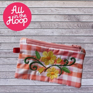 Peut inclure: Une pochette zippée en tissu vichy rose et blanc avec des fleurs jaunes et des feuilles vertes brodées. La pochette est sur un fond en bois.