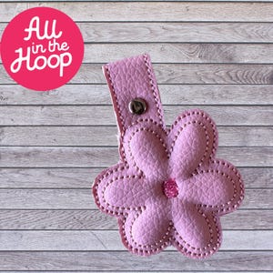 May include: Pink flower-shaped key chain with a metal loop and a silver button. The keychain is made of faux leather and has a stitched design.