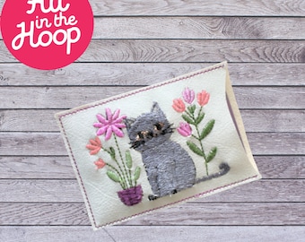 May include: A white fabric pouch with a gray cat and pink flowers embroidered on it. The pouch is on a wooden background.