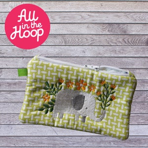 Peut inclure: Une pochette zippée verte avec une fermeture éclair blanche et un éléphant brodé blanc avec des fleurs sur un fond vert. La pochette a une boucle en tissu vert.