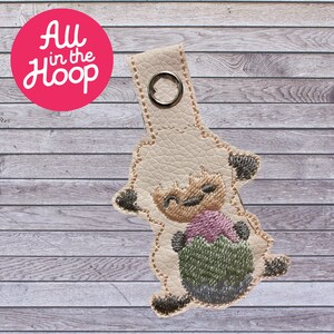 May include: A white lamb with a pink and green Easter egg. The lamb is holding the egg in its front paws. The lamb is embroidered on a beige leather keychain.