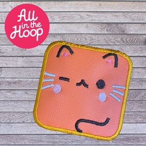May include: A peach-colored felt cat face with black eyes, a pink nose, and a black tail. The cat face is surrounded by a gold-colored border.