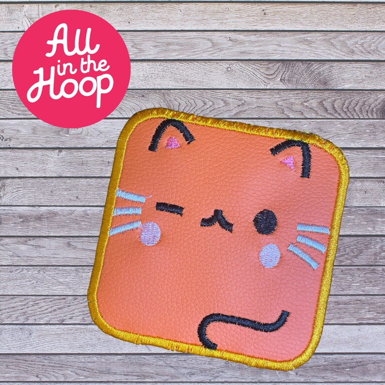 May include: A peach-colored felt cat face with black eyes, a pink nose, and a black tail. The cat face is surrounded by a gold-colored border.