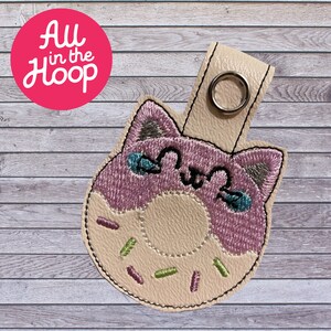 May include: A pink and white donut with a cat face embroidered on it. The donut has sprinkles and a hole in the middle. The donut is attached to a key chain.