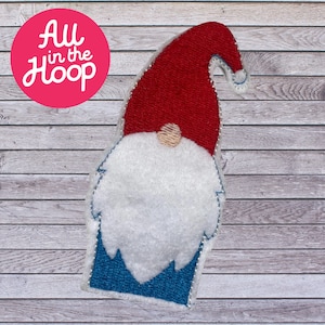 Peut inclure: Un gnome en feutre rouge et blanc avec un chapeau bleu et une barbe blanche. Le gnome est debout sur un fond en bois.