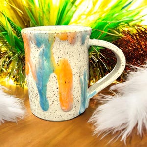 May include: A white ceramic mug with a speckled glaze and abstract blue, orange, and yellow drips. The mug has a rounded handle.